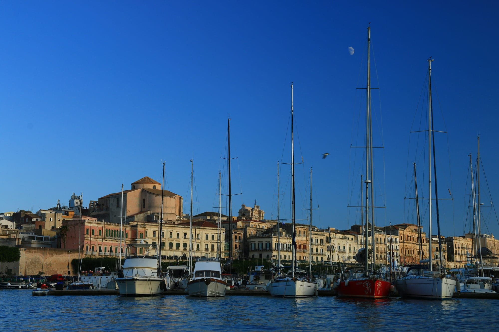 Ortigia seen from the water: moored boats, the seafront palazzi and a dome behind them