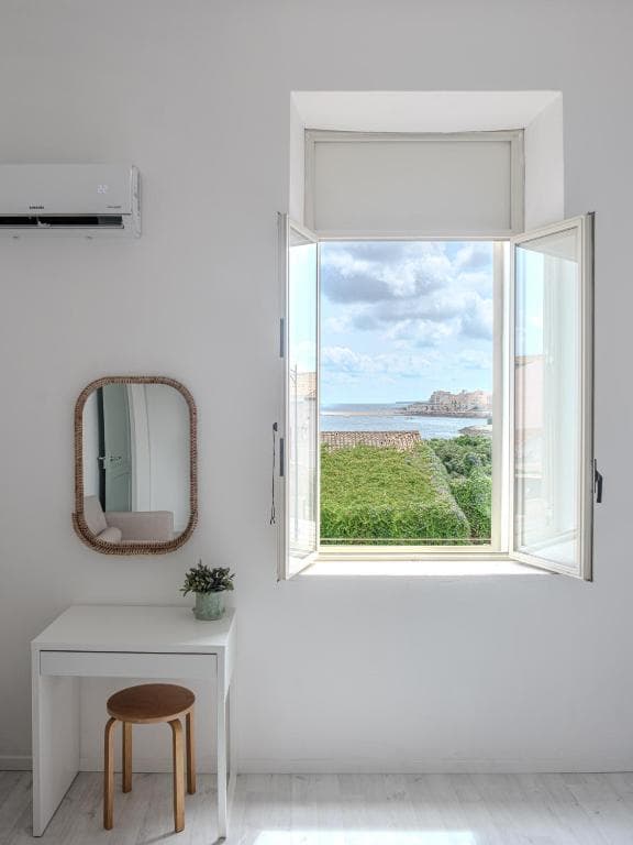 An open bedroom window onto the sea: sky, water and the headland on the horizon, with a mirror and a small white table beside it