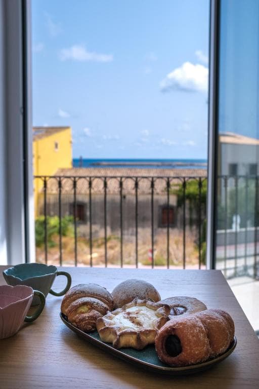 Breakfast on the table by the open window: pastries on a tray, two cups, and the sea visible past the railing