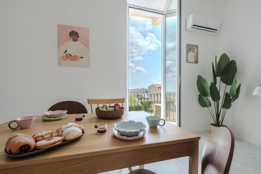 The dining table laid for breakfast in front of the open balcony door onto the sea, with the plant and the pictures on the walls